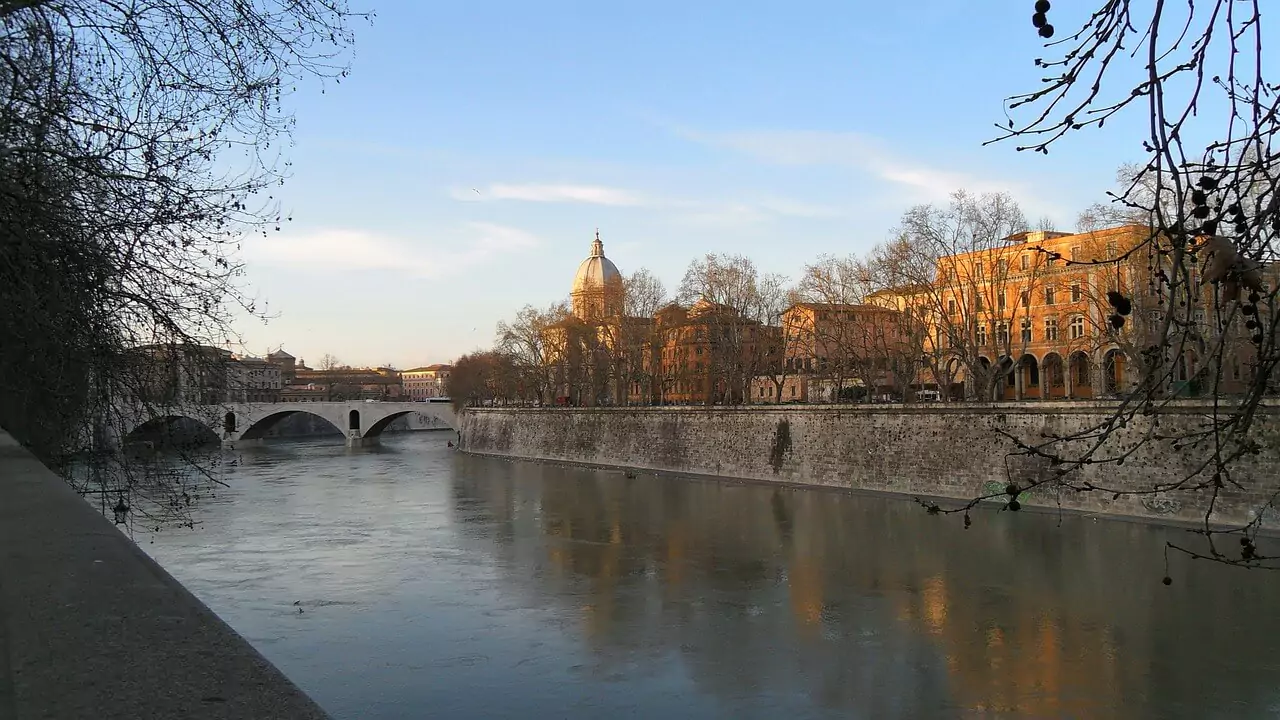 View of the river in Trastevere, Rome, with calm waters reflecting the historic bridges, leafy trees lining the banks, and the charming architecture of the lively neighborhood in the background.