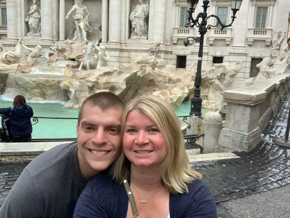 Katie and Zack standing in front of the Trevi Fountain in Rome during the morning, smiling with the stunning Baroque sculptures and cascading water behind them. The soft morning light highlights the fountain’s intricate details and adds a peaceful glow to the scene.