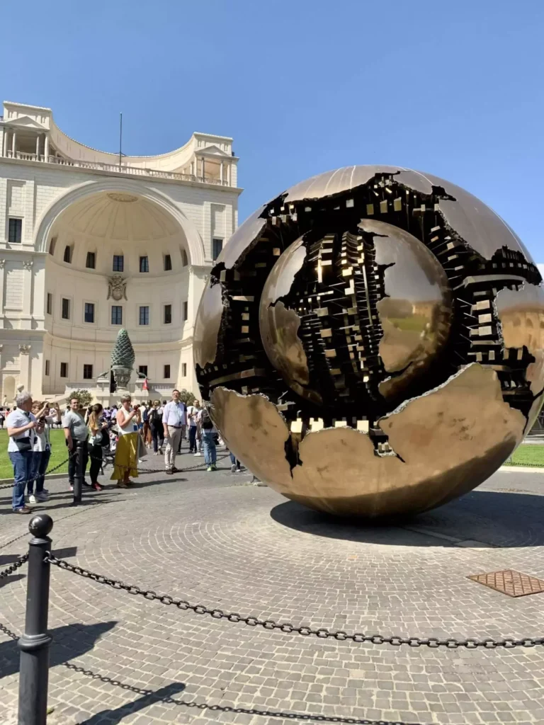 Sphere Within a Sphere by Arnaldo Pomodoro at the Vatican Museums in Rome, featuring a large bronze globe with a fractured, mechanical inner sphere exposed. The modern sculpture contrasts with the historic setting, symbolizing complexity within harmony.