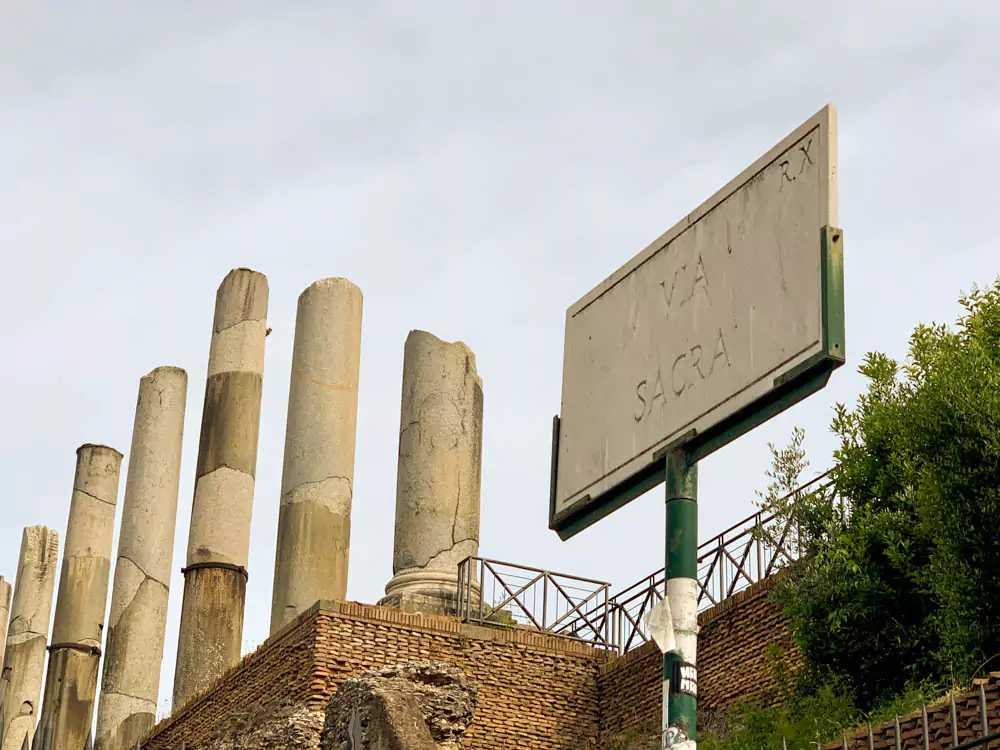 Sign for the Via Sacra in the Roman Forum, with the ruins of the Temple of Venus and Roma rising in the background, featuring ancient columns and arches under a sunny Roman sky.