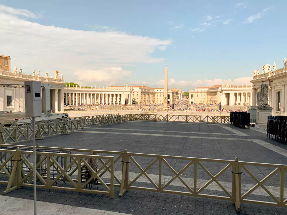 The sun casting long shadows across St. Peter’s Square in Vatican City, highlighting the colonnades, cobblestones, and visitors walking through the iconic open plaza in the soft afternoon light.