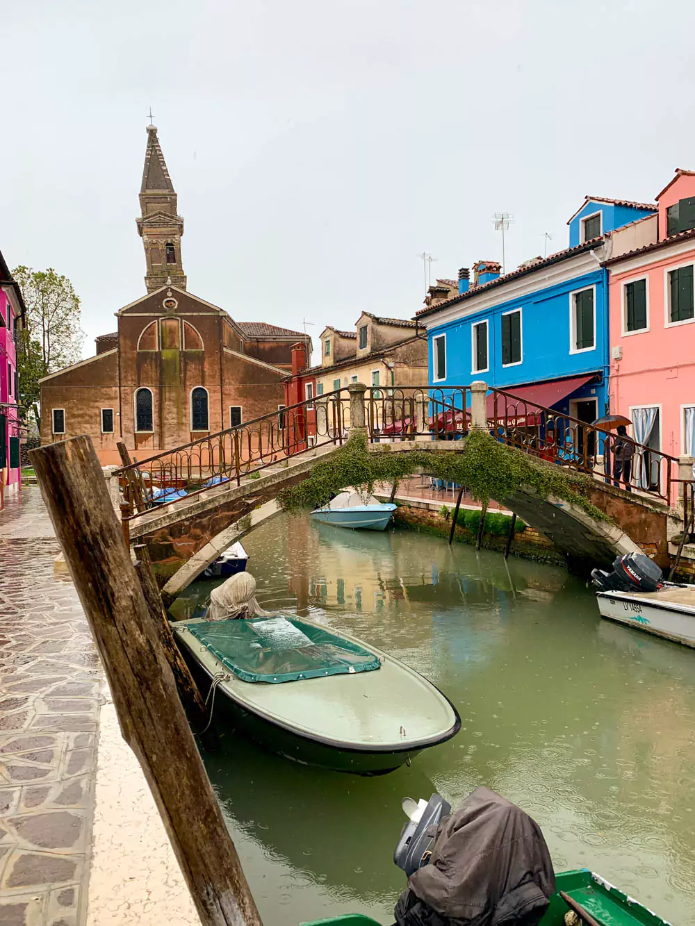 The main canal of Burano, Italy, on a rainy day, with reflections of brightly painted houses shimmering in the wet pavement and canal water, as small boats line the water’s edge under a gray sky.