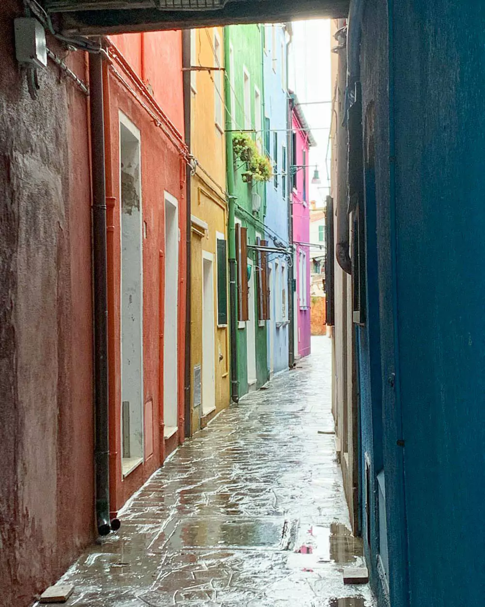 Narrow alley in Burano, Venice, lined with brightly colored houses in shades of pink, blue, yellow, and green, creating a cheerful and picturesque walkway through the island village.