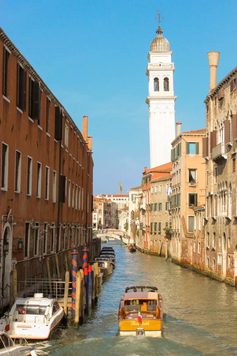 Quiet canal scene near the San Giorgio dei Greci church in Venice, Italy, with a gently leaning bell tower, pastel-colored buildings, and a small arched bridge reflecting in the still water.