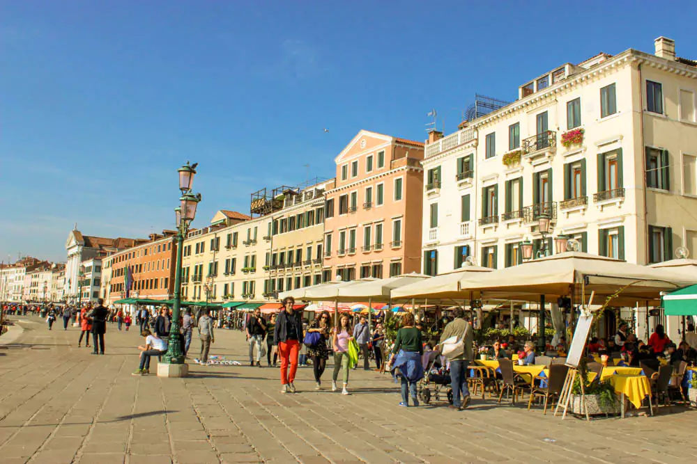 Venice’s Riva degli Schiavoni waterfront promenade, bustling with tourists, lined with historic buildings, market stalls, and views of gondolas and boats along the sparkling lagoon.