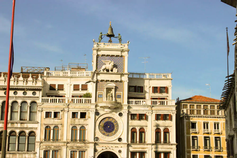 The Clock Tower in St. Mark’s Square, Venice, Italy, featuring its ornate blue and gold astronomical clock, arched passageway below, and statues atop the structure, set against historic Venetian buildings.