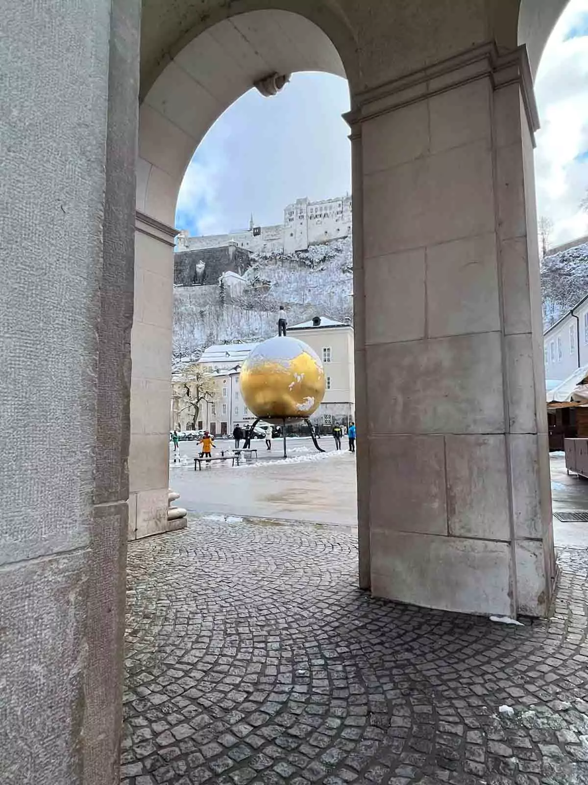 Kapitelplatz in Salzburg, Austria, with the Goldene Kugel sculpture—a large golden sphere topped with a standing male figure—set against views of Salzburg Cathedral and the Hohensalzburg Fortress.