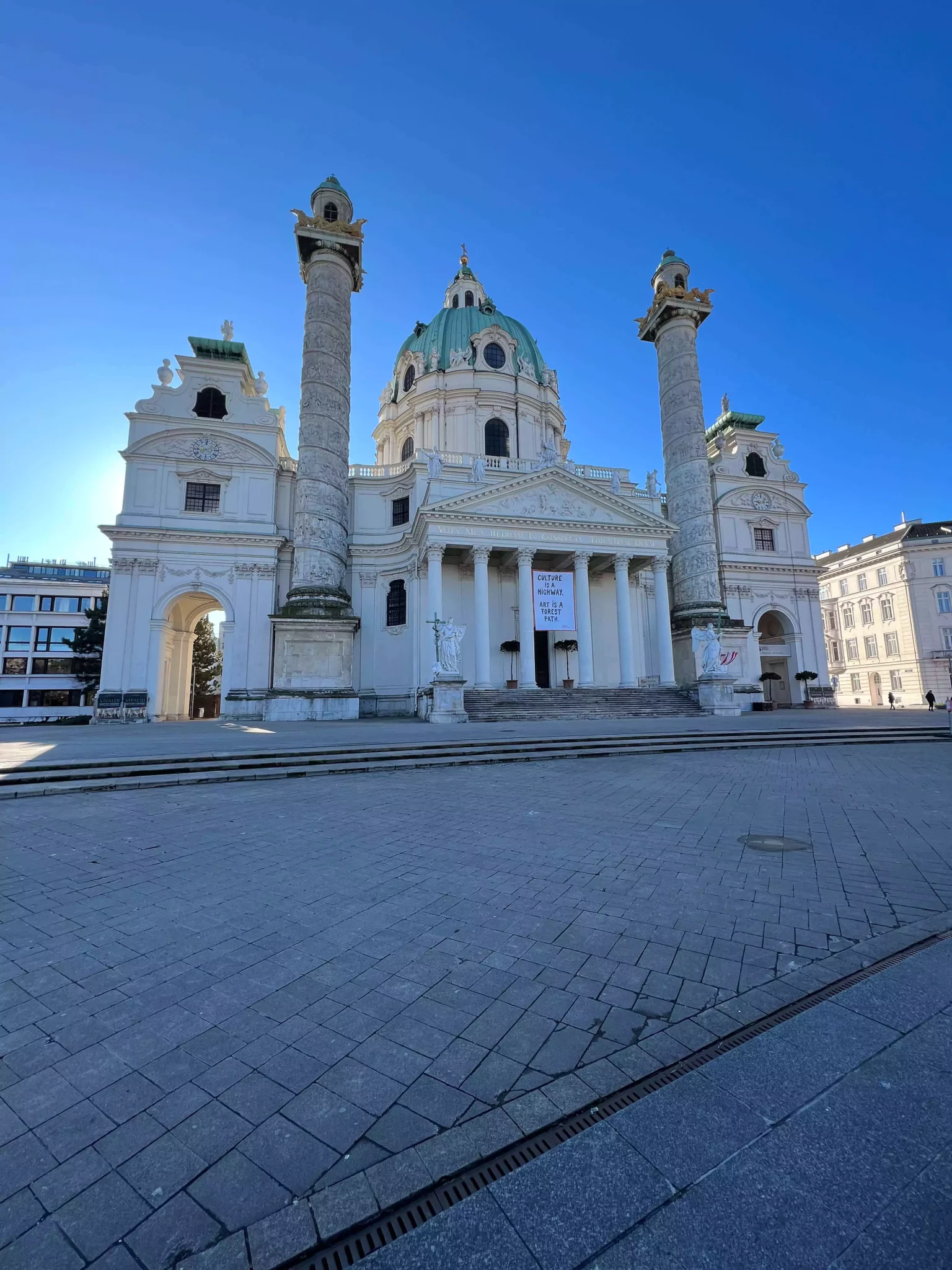 The exterior of Karlskirche in Vienna, Austria, stands beautifully lit under a clear, sunny sky with no people in sight. Its grand dome, twin columns, and baroque façade shine against the vivid blue backdrop, highlighting the church’s architectural elegance.