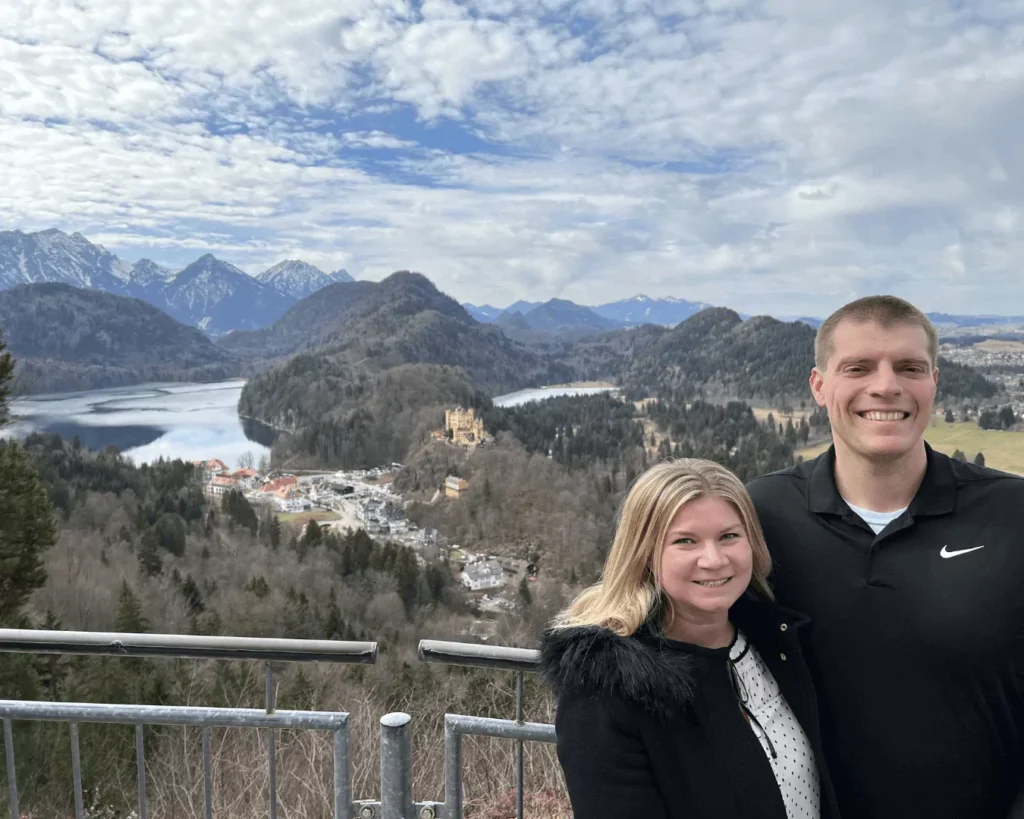 Katie and Zack standing at a scenic viewpoint above Hohenschwangau Valley in Bavaria, Germany, with sweeping views of forested hills, winding roads, and alpine lakes stretching below.