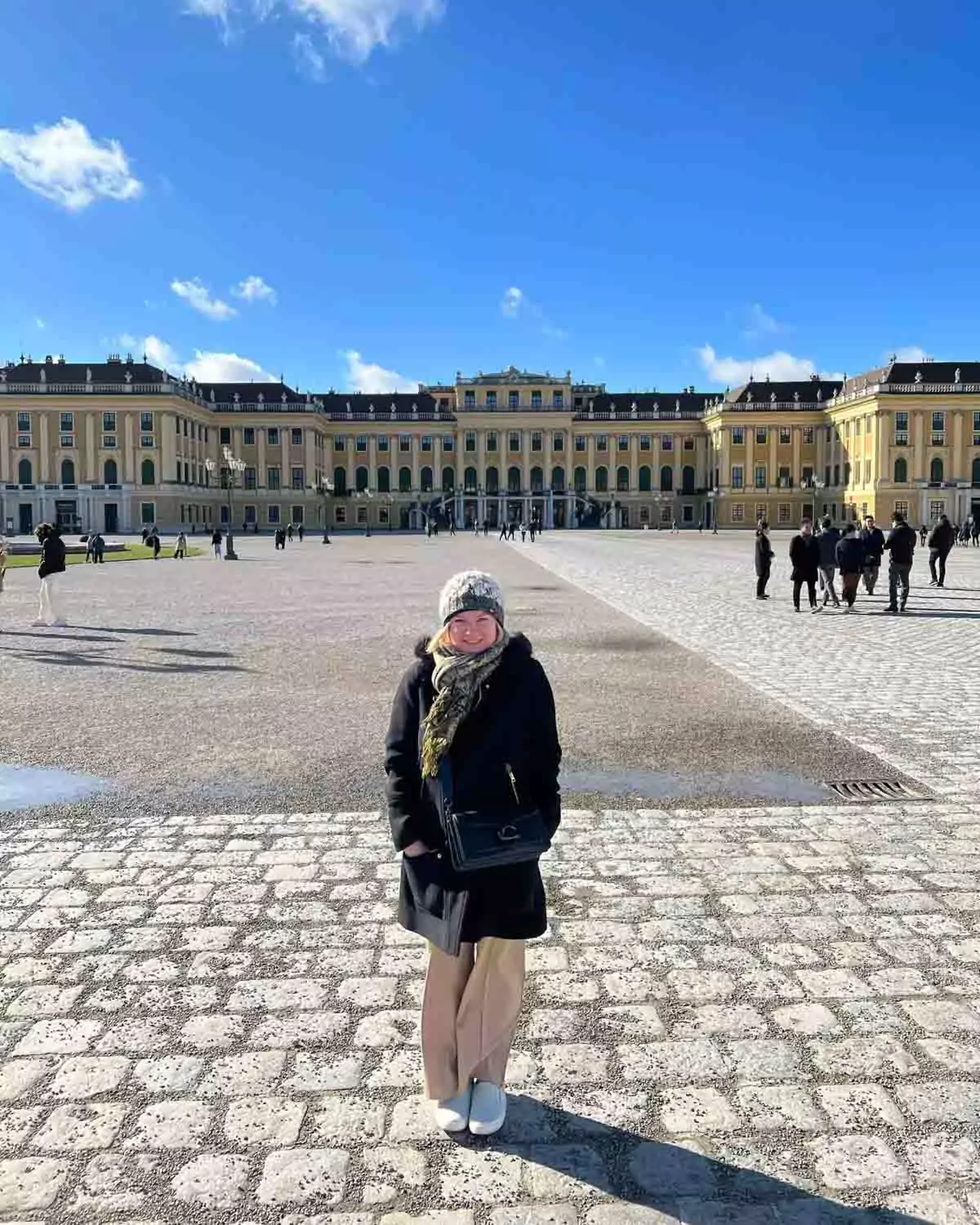 Katie standing at the entrance of Schloss Schönbrunn in Vienna, Austria, with the grand yellow baroque palace and ornate gates behind her on a partly cloudy day.