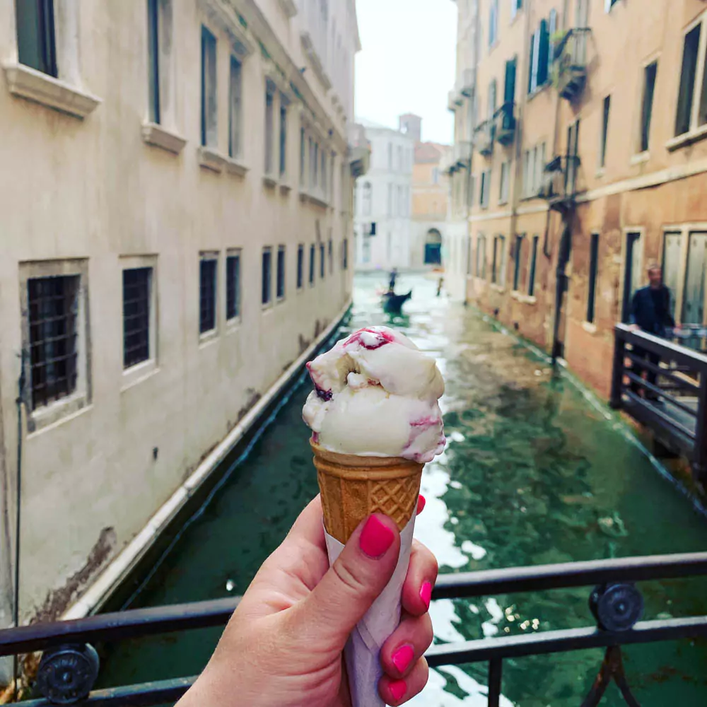 Katie holding a cone of cherry gelato above a canal in Venice, Italy, with the vibrant red scoop in focus and charming Venetian buildings and a narrow waterway in the background.