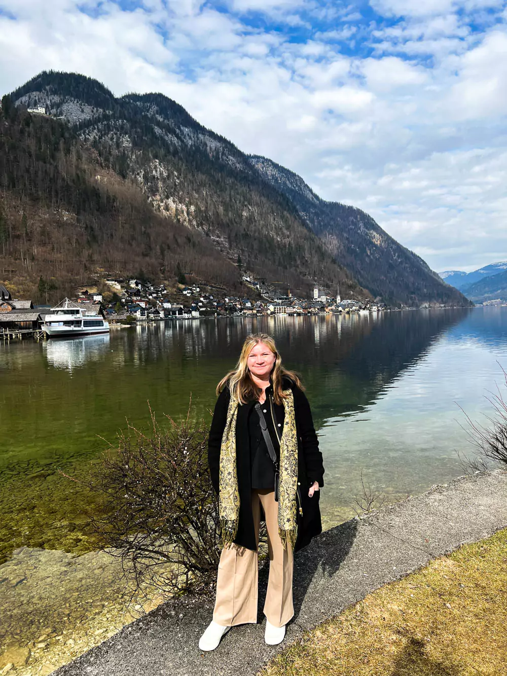 Katie standing by the edge of the water in Hallstatt, Austria, with the charming village and its historic buildings reflected across the pond under a peaceful, cloudy sky.