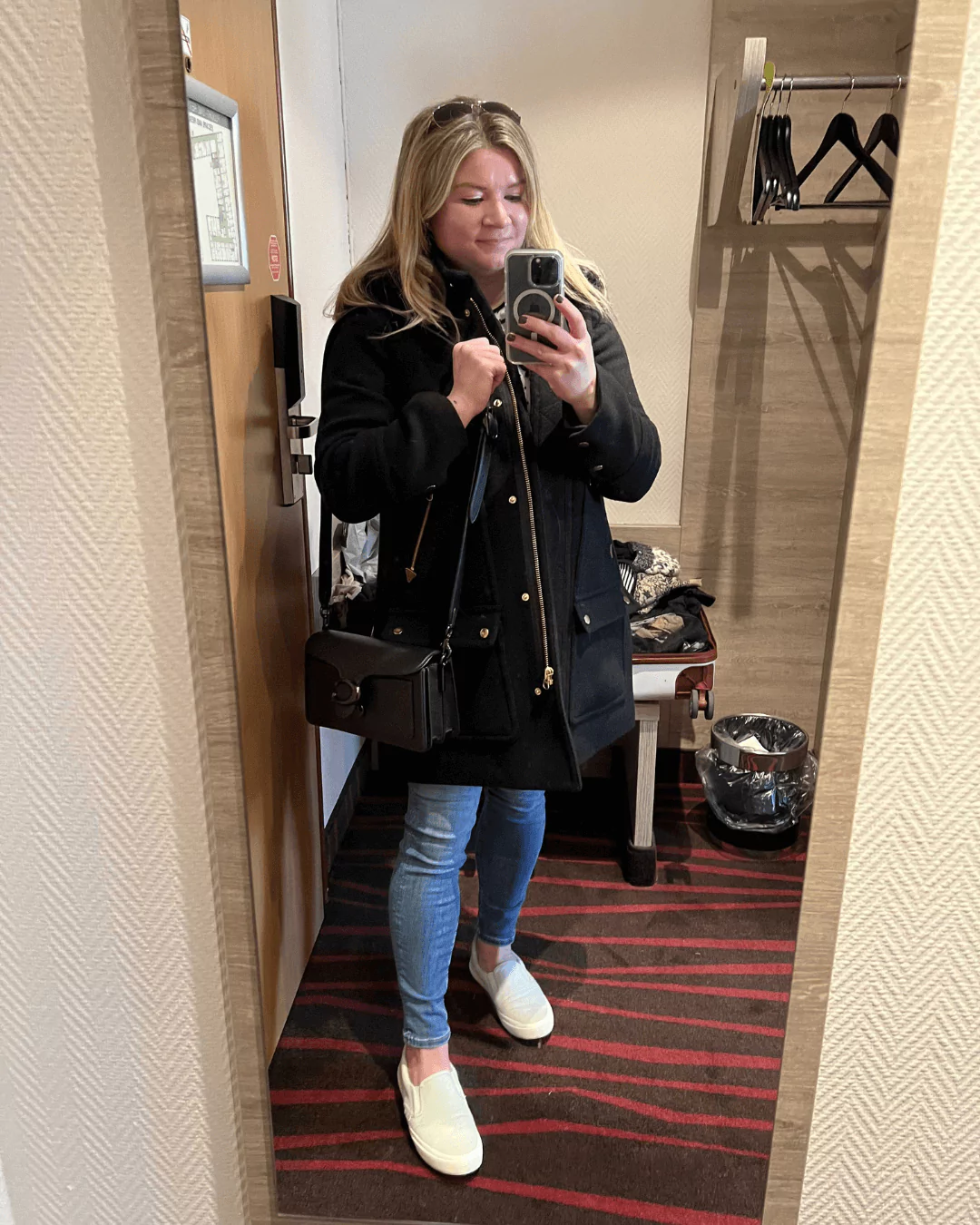 Katie taking a mirror selfie in Munich, Germany, wearing a black jacket and casual travel attire, with a softly lit interior space reflected in the background.