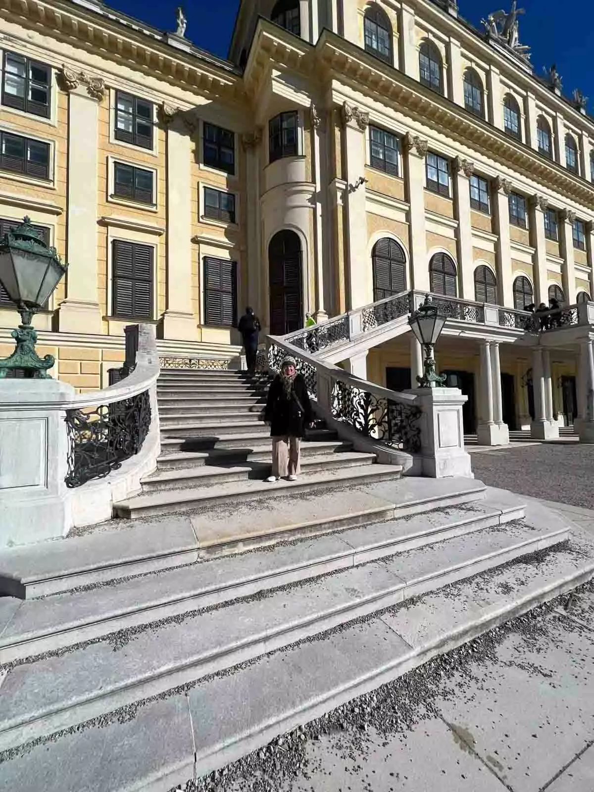 Katie standing on the grand stone staircase at Schloss Schönbrunn Palace in Vienna, Austria, with the Baroque palace’s yellow facade and elegant architectural details in the background.