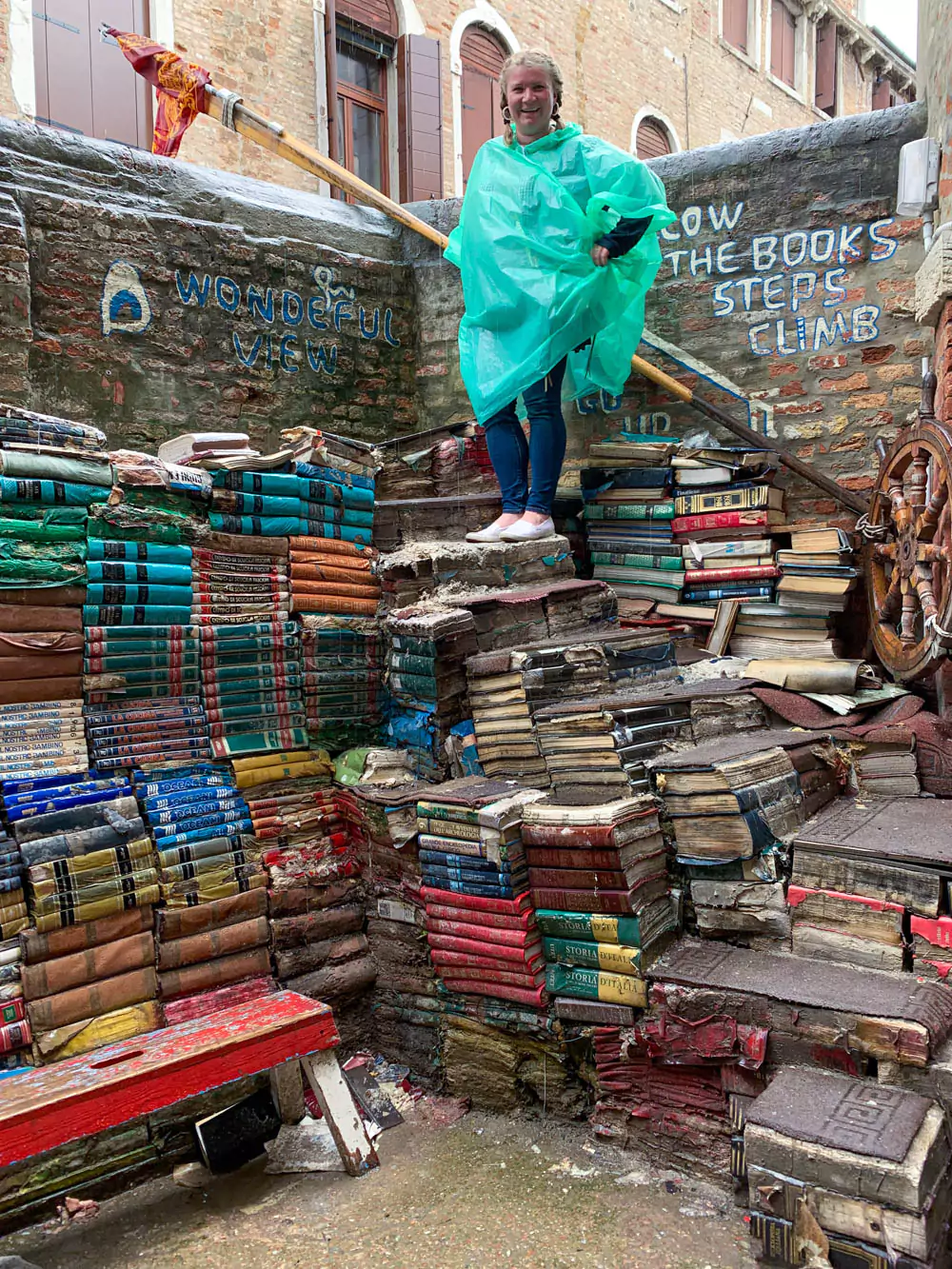 Katie standing on the iconic book staircase at Libreria Acqua Alta in Venice, Italy, surrounded by weathered books stacked to form steps, with a brick wall and canal view in the background.