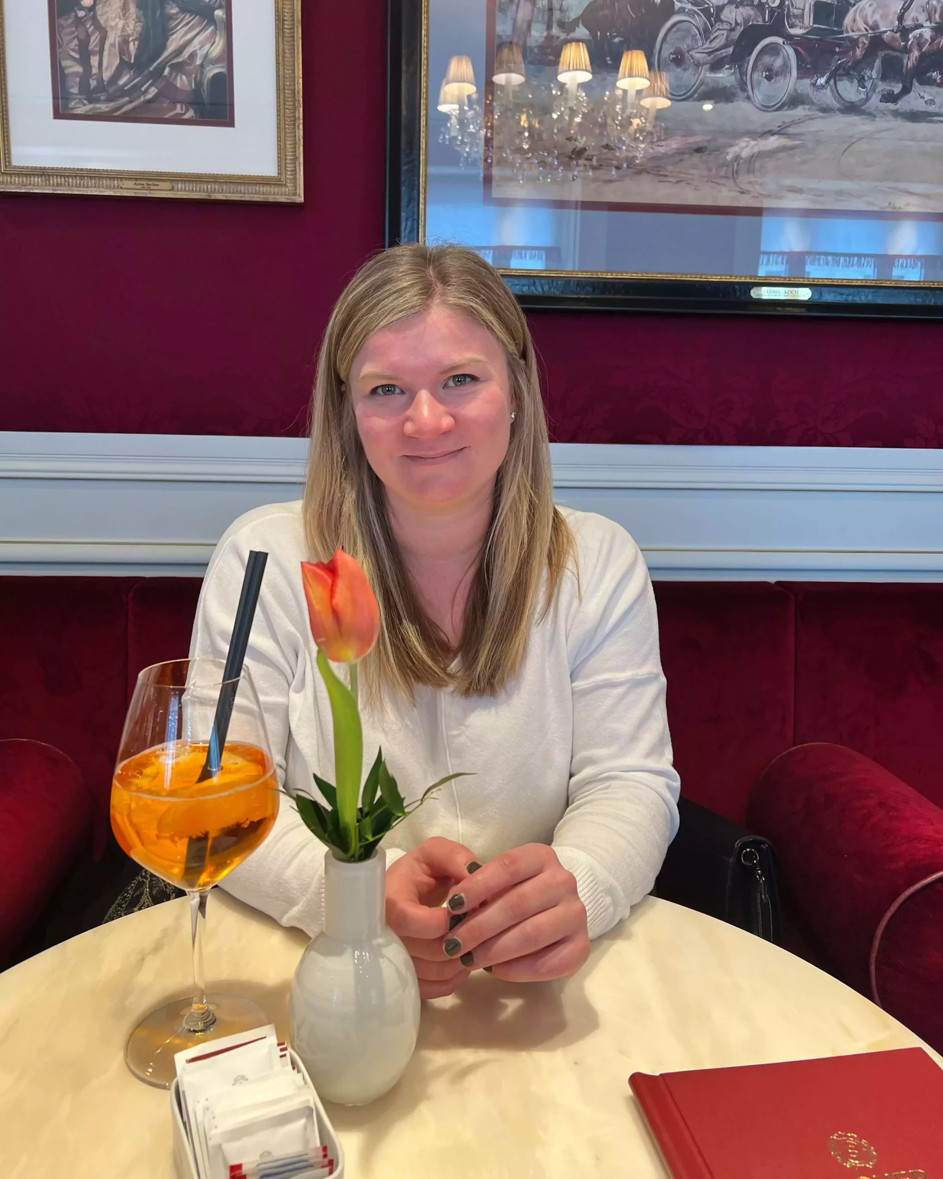 Katie sits at a small outdoor table at Café Sacher in Salzburg, Austria, enjoying an Aperol Spritz. The elegant setting and her relaxed pose capture a perfect moment of European café culture.