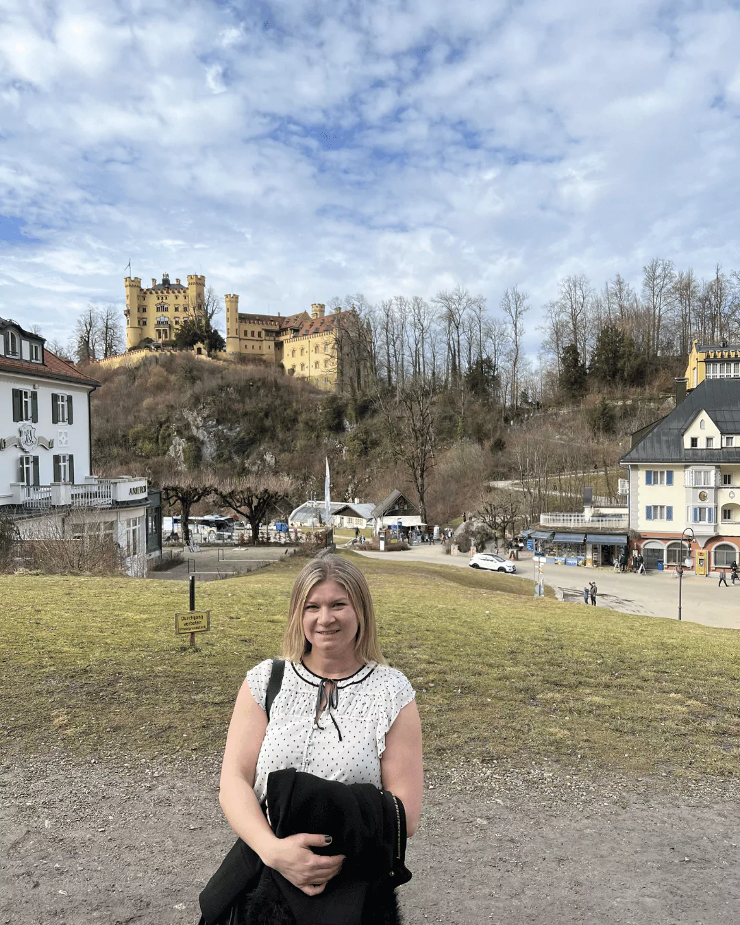 Katie standing in Schwangau Village, Germany, with the yellow towers of Hohenschwangau Castle visible on the wooded hillside behind her, surrounded by alpine scenery and traditional Bavarian charm.