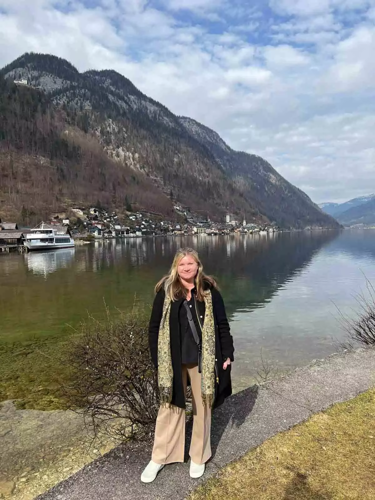 Katie standing next to the Hallstätter See in Hallstatt, Austria, with calm lake waters, alpine mountain reflections, and the charming lakeside village in the background.