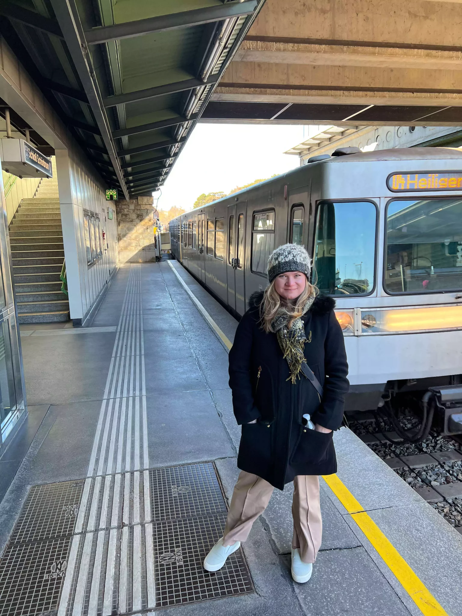 Katie stands on the Schönbrunn station platform in Vienna, Austria, with the historic station’s yellow walls and arched windows behind her. The quiet platform and classic design reflect the charm of Vienna’s transit system near the famous palace.