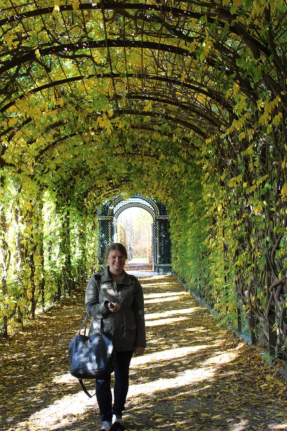 Katie stands beneath a lush green arched trellis at Schönbrunn Palace in Vienna, Austria. The leafy tunnel casts soft shadows along the gravel path, creating a serene, fairytale-like moment in the palace gardens.