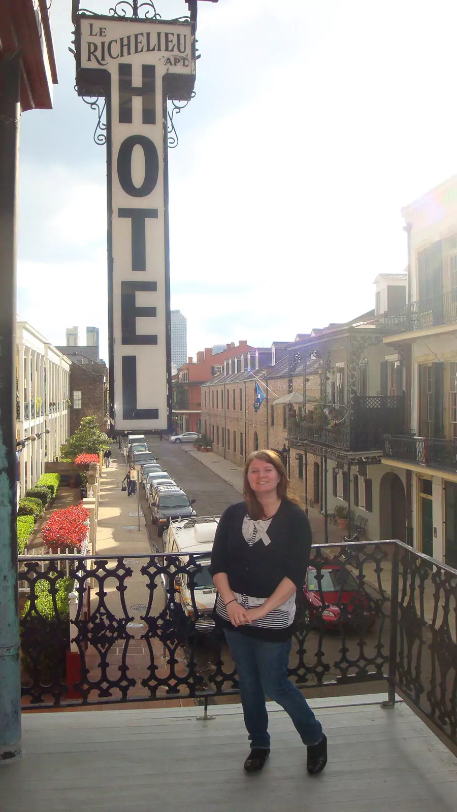 Katie stands smiling on the ornate balcony of Hotel Le Richelieu in New Orleans, Louisiana, with intricate wrought-iron railings and colorful historic buildings in the background. The scene captures the charm of the city’s French Quarter architecture.