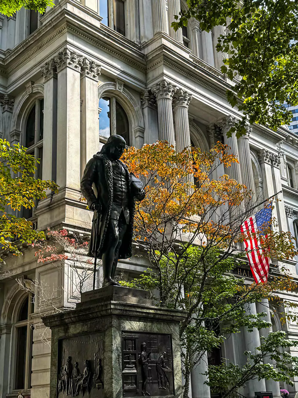 Statue of Benjamin Franklin in front of Boston City Hall, standing on a stone pedestal with engraved text. The historic figure is depicted in colonial attire, set against the backdrop of modern architecture and city life.