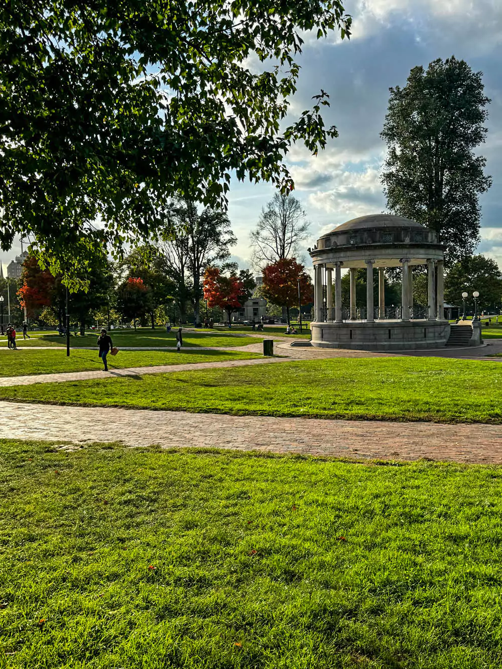 A white wooden gazebo with a pitched roof stands in Boston Common, surrounded by green grass and mature trees. The classic structure serves as a charming focal point in the city’s historic public park."