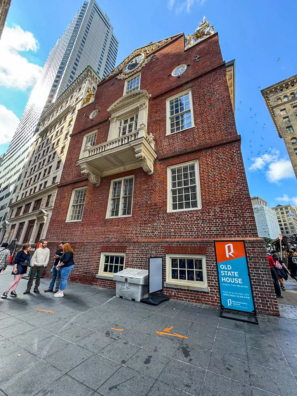 Brick Old State House in Boston with white trim, a prominent balcony, and a golden lion and unicorn atop the roof. Set against modern skyscrapers, this colonial-era building marks the site of key events like the Boston Massacre and stands as a symbol of American independence.