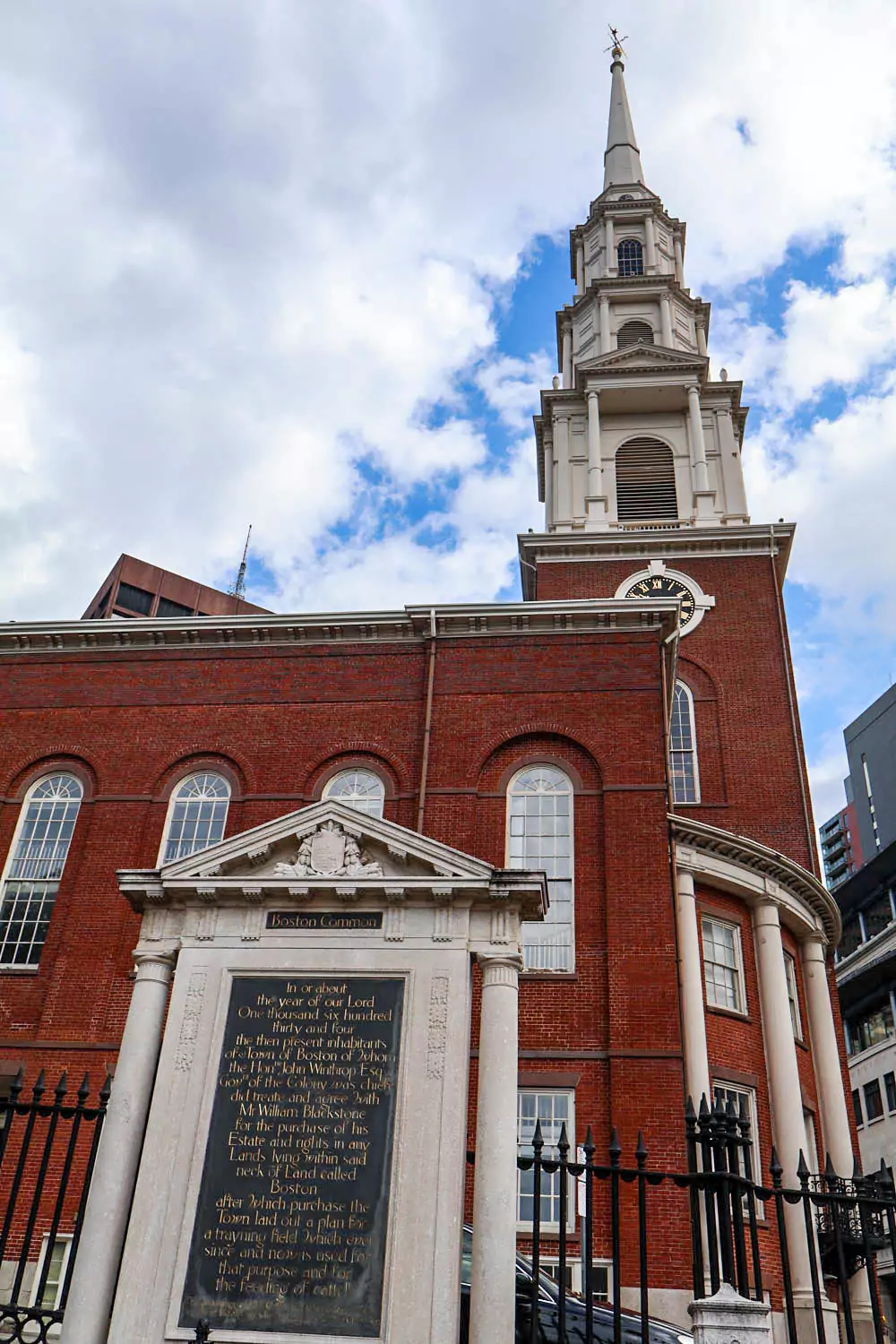 The white steepled Park Street Church stands tall against a clear blue sky in downtown Boston, featuring classic colonial architecture with large columns and arched windows. The historic church is a prominent landmark near Boston Common.