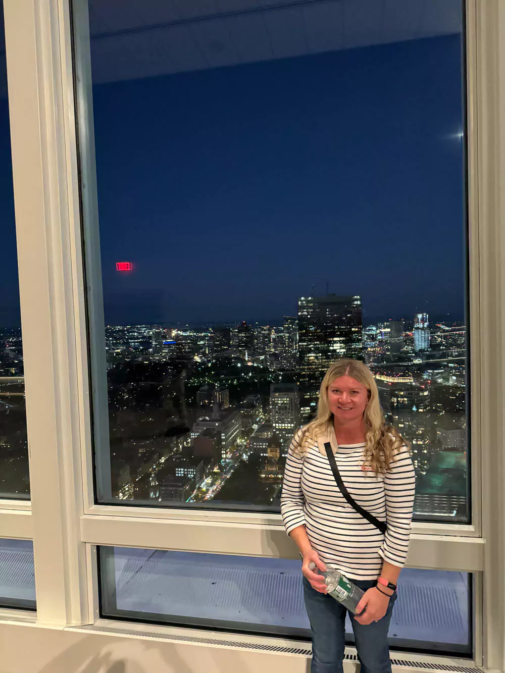 Katie standing in front of the Boston skyline at night, with city lights reflecting off the water and skyscrapers glowing against the dark sky. The illuminated backdrop highlights the city’s vibrant and modern charm.