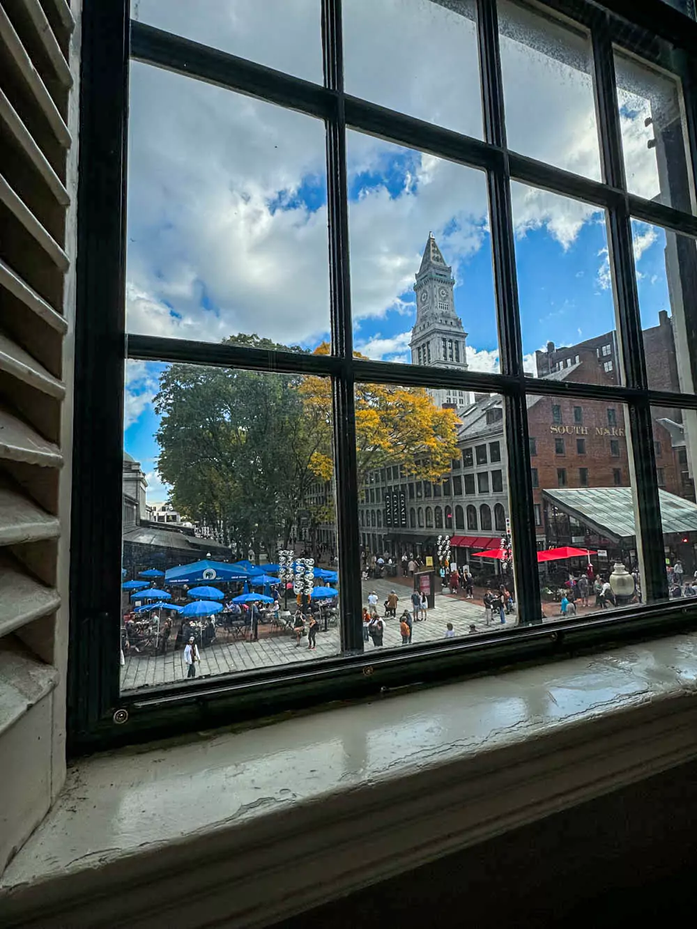 Looking toward South Market from Faneuil Hall in Boston, the bustling marketplace features historic brick buildings, outdoor dining areas, and shoppers walking along the streets. The scene captures the vibrant atmosphere of this popular urban shopping and dining district.