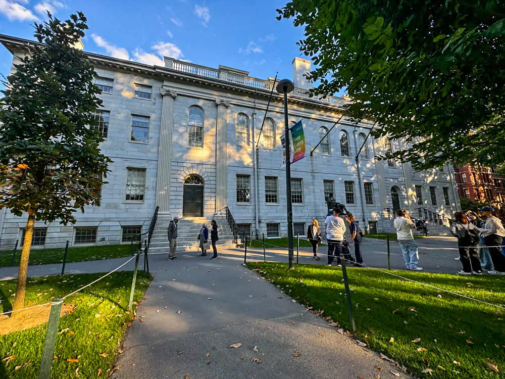 University Hall at Harvard University, a stately red-brick building with white columns and a pitched roof, set amidst green lawns and trees on campus. The neoclassical architecture reflects the historic academic environment.