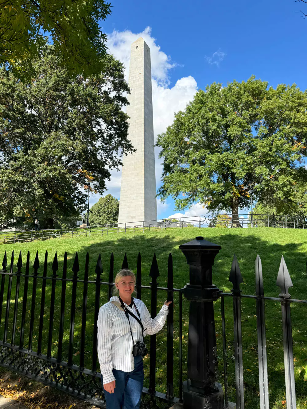 Katie standing in front of the gates of the Bunker Hill Memorial in Charlestown, Massachusetts, with the tall granite obelisk visible behind her. The historic monument honors a pivotal battle of the American Revolution.