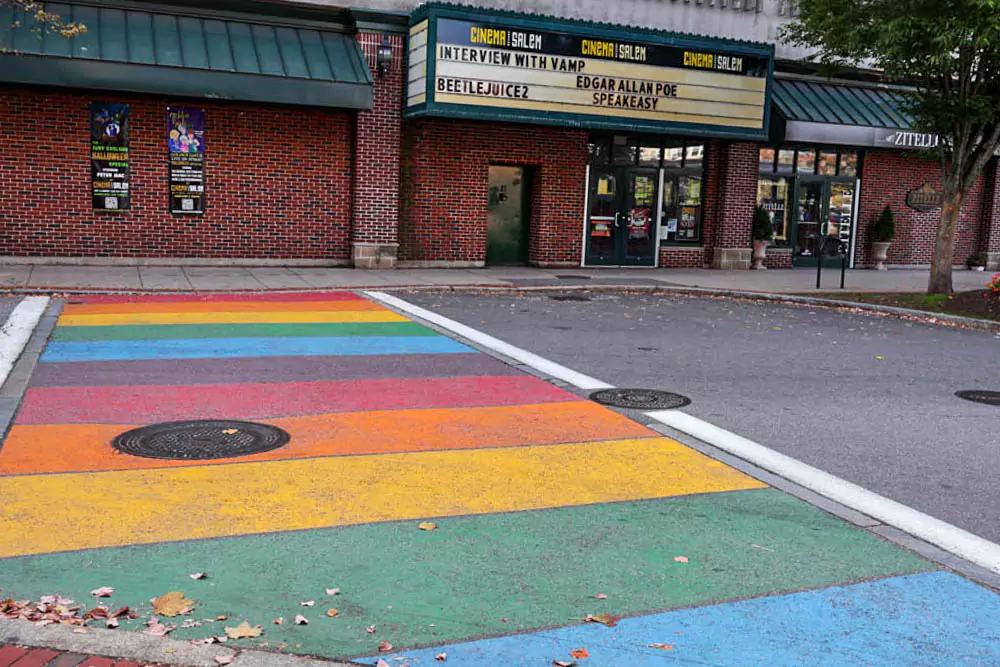 Movie theatre sign in Salem, Massachusetts, with retro-style lettering above a rainbow-painted crosswalk. The colorful scene blends local pride with vintage charm in the heart of the city.