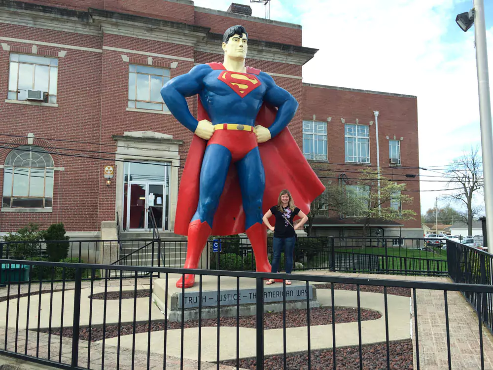 Katie stands smiling beside the large Superman statue in Metropolis, Illinois, known as the home of Superman. The colorful statue depicts the superhero in his classic costume with city buildings visible in the background.