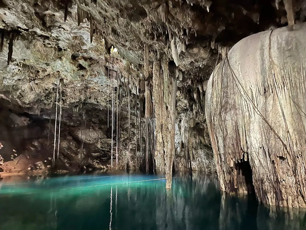 Cenote Xkekén in Mexico, an underground cave cenote with clear blue water, dramatic stalactites hanging from the ceiling, and soft light filtering through a small natural opening above.