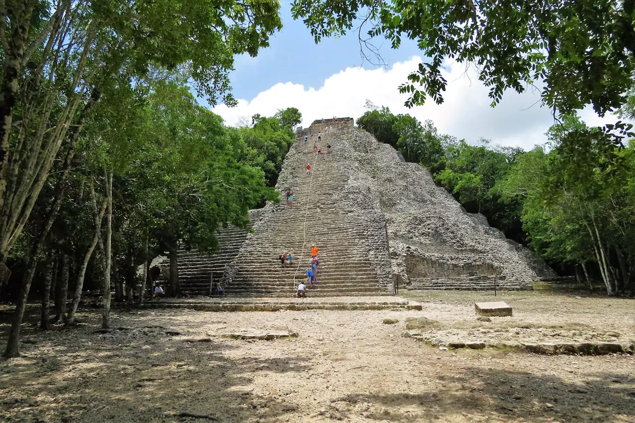 A tall stone pyramid with many steps surrounded by trees, with people climbing it.