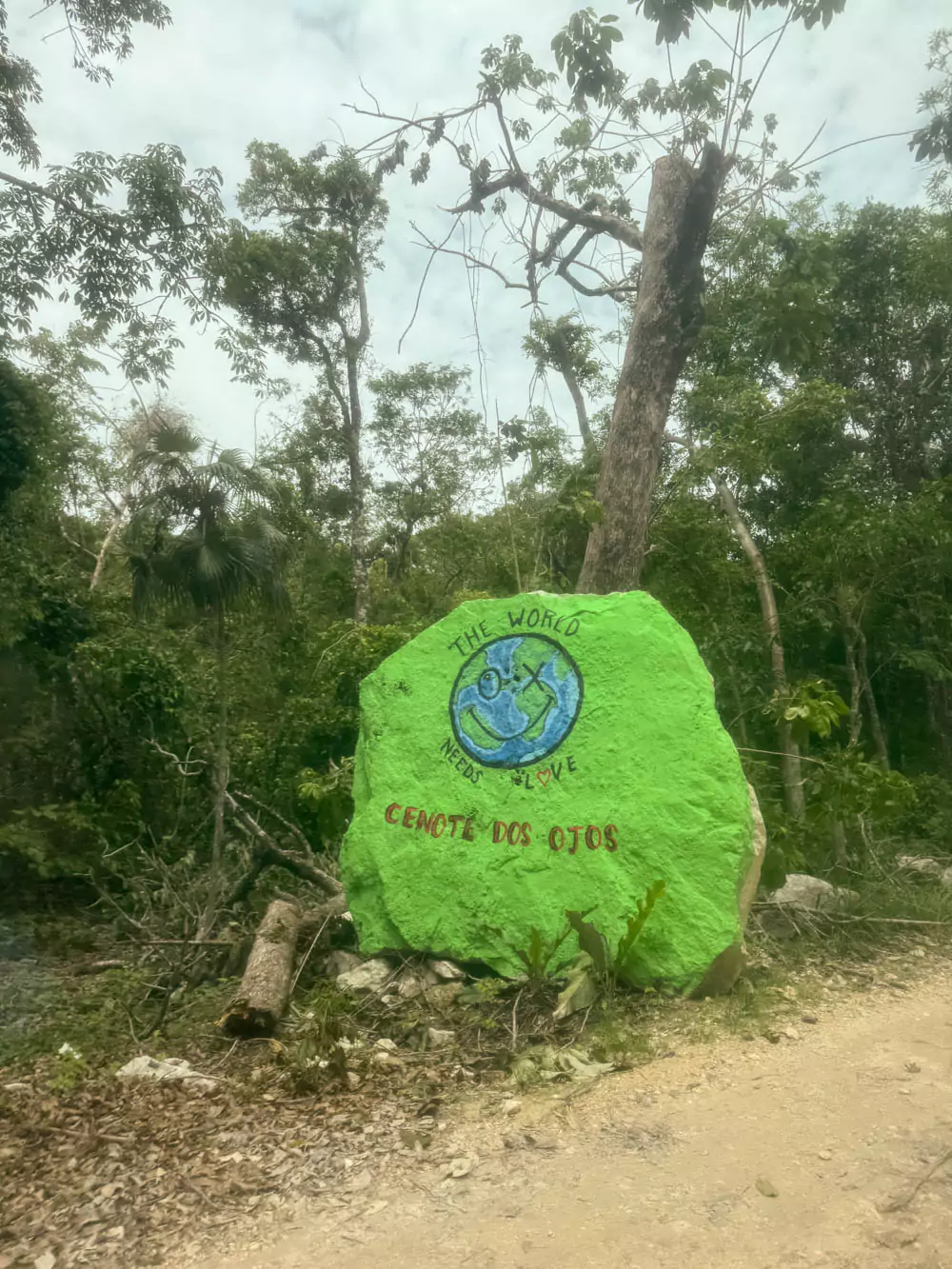 Painted green rock marking the entrance to Cenote Dos Ojos in Mexico, with the name “Dos Ojos” in bold white lettering, surrounded by tropical plants and jungle scenery.