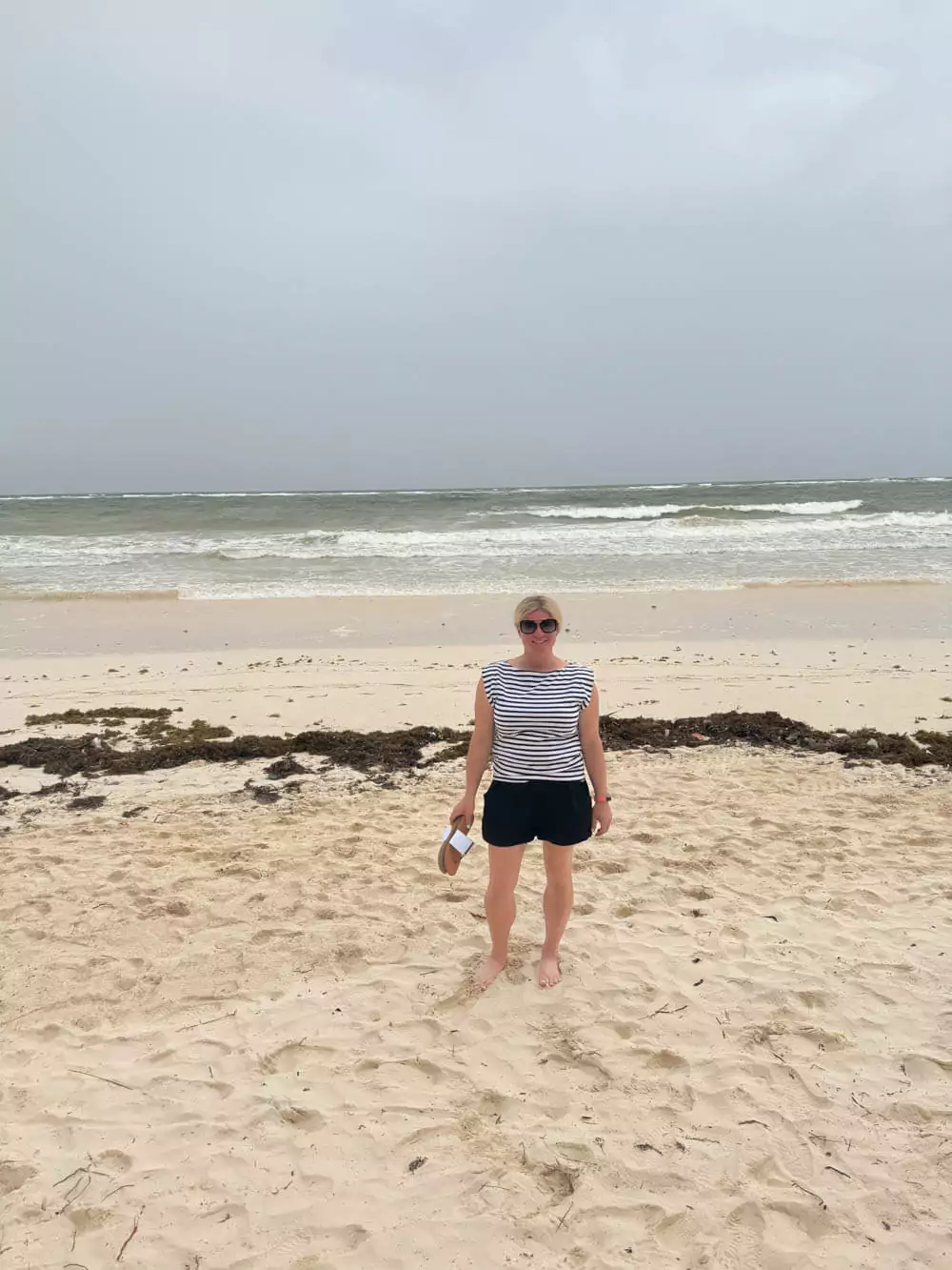 Katie standing on a Tulum beach with patches of seaweed along the shoreline, looking out at the turquoise water under a partly cloudy sky.
