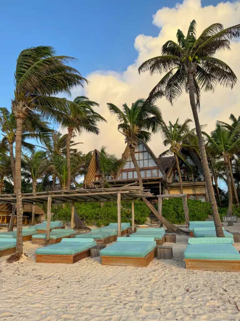 Rows of comfortable beach beds with white cushions arranged on the sandy shore in front of La Valise Tulum’s main building, framed by tropical greenery. The inviting setup overlooks the turquoise ocean, perfect for relaxing by the water.