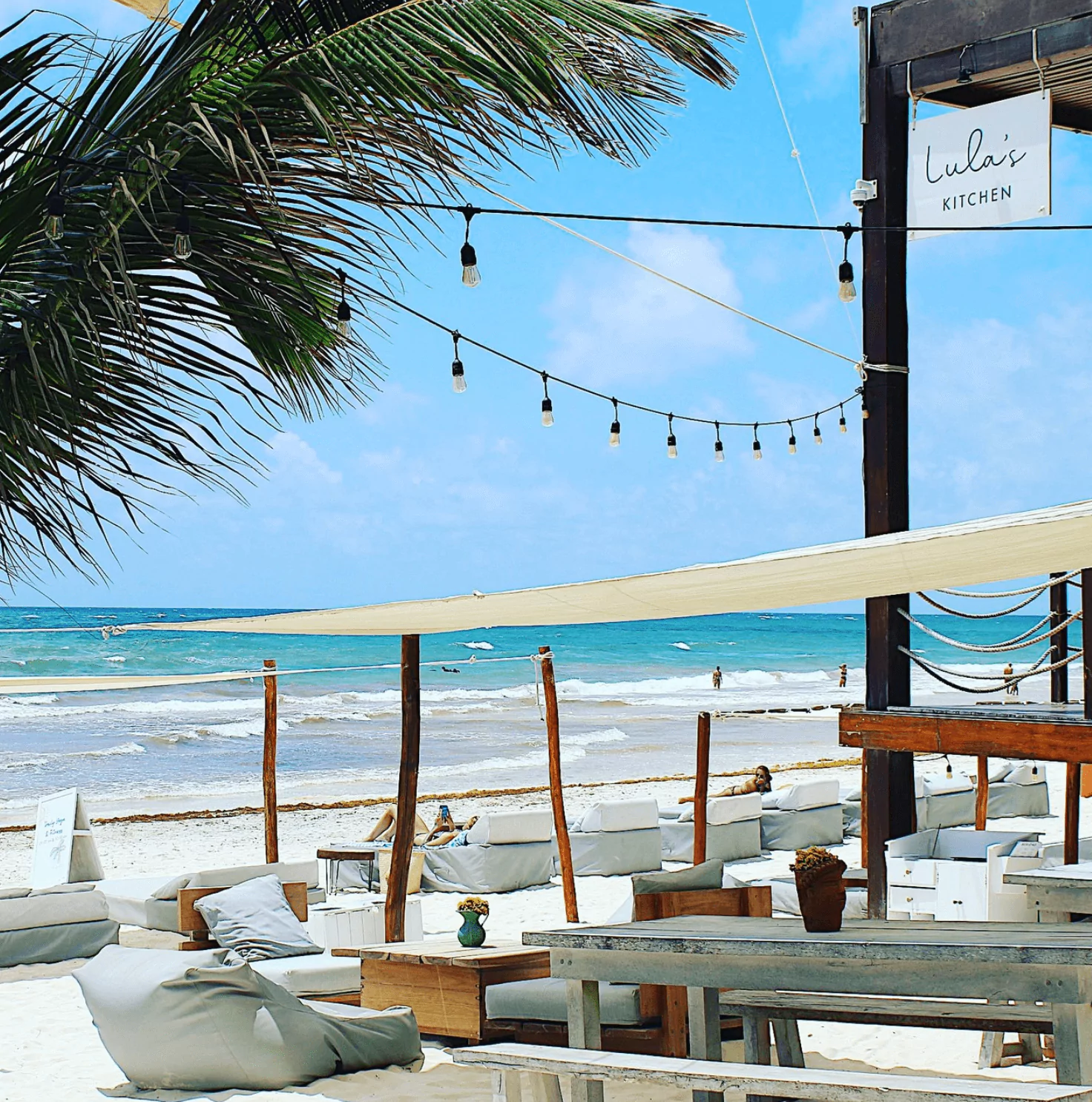 Beachfront dining setup at Lula Hotel in Tulum, featuring rustic wooden tables, woven chairs, and ocean views framed by palm trees and soft Caribbean breezes.