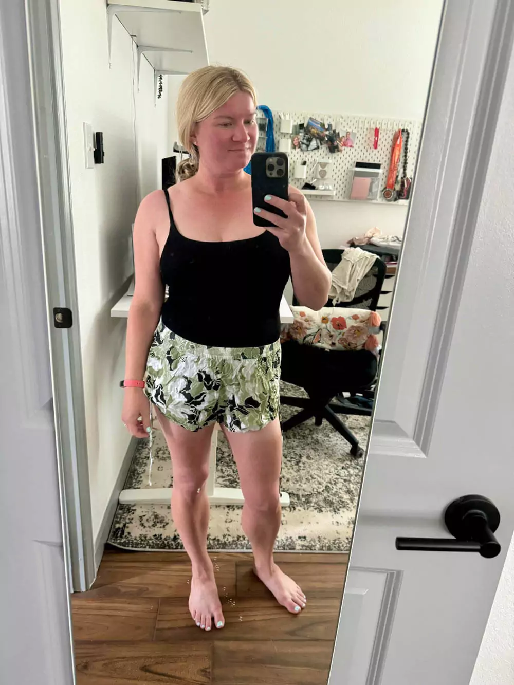 Katie wearing a black one-piece swimsuit paired with palm print shorts, front view, standing in a tropical setting with relaxed beach vibes.