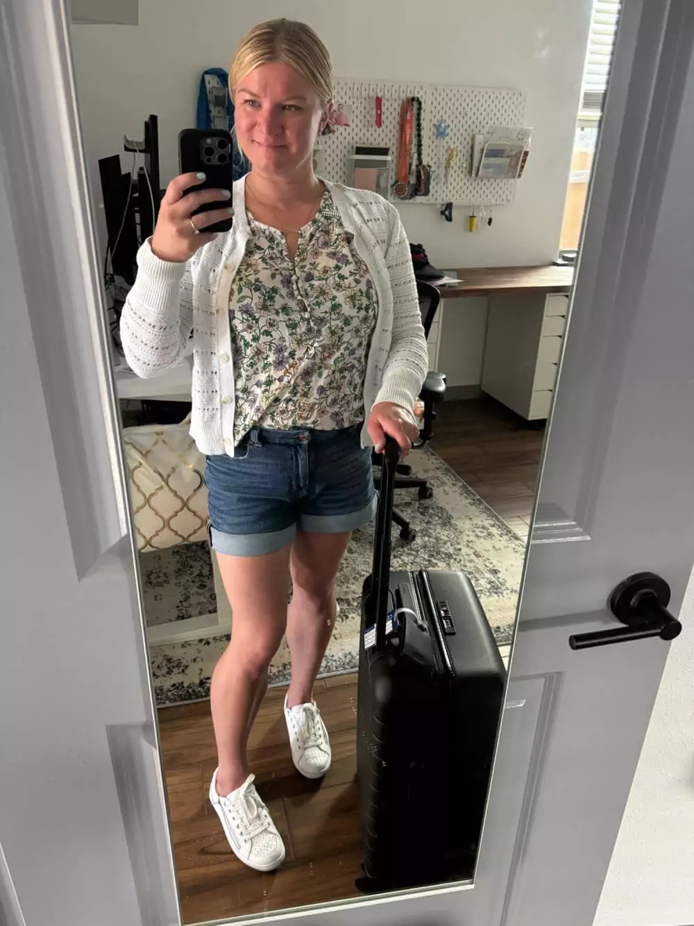 Travel outfit with jean shorts, floral top, cardigan, sneakers, earrings, and carry-on suitcase.