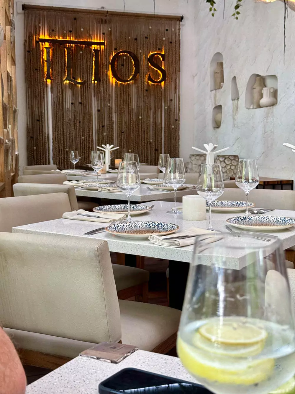 Elegant dining tables at Ilios Tulum, set with sleek tableware and surrounded by stylish boho-Greek decor, warm lighting, and a vibrant, upscale atmosphere.