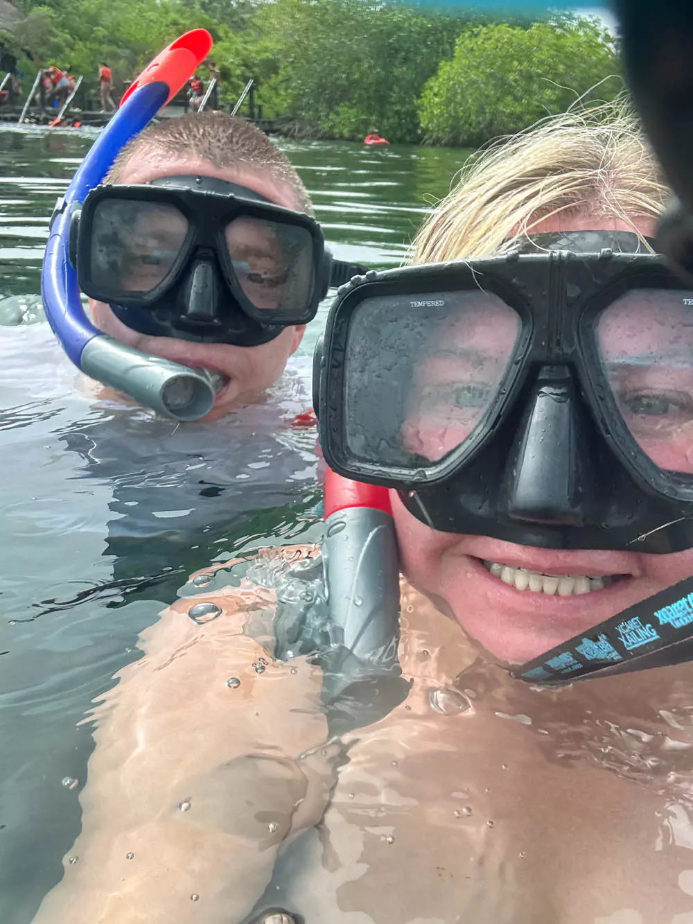 Katie and Zack snorkeling in the crystal-clear waters of Yal-Ku Lagoon in Mexico, surrounded by vibrant tropical fish and rocky underwater formations in a calm, sunny setting.