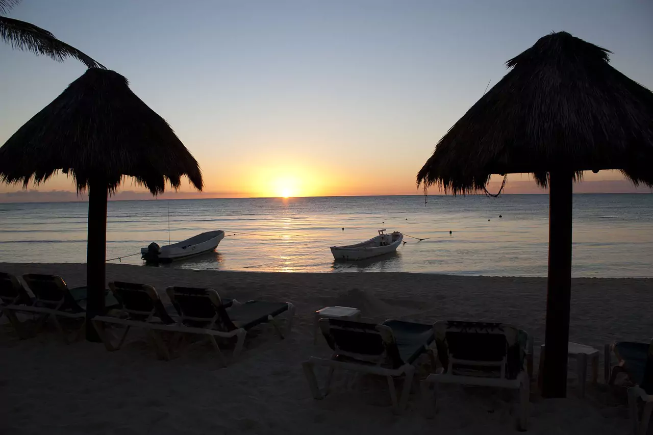 A sunset over calm ocean waters with two boats anchored near a sandy beach and palm-thatched umbrellas.