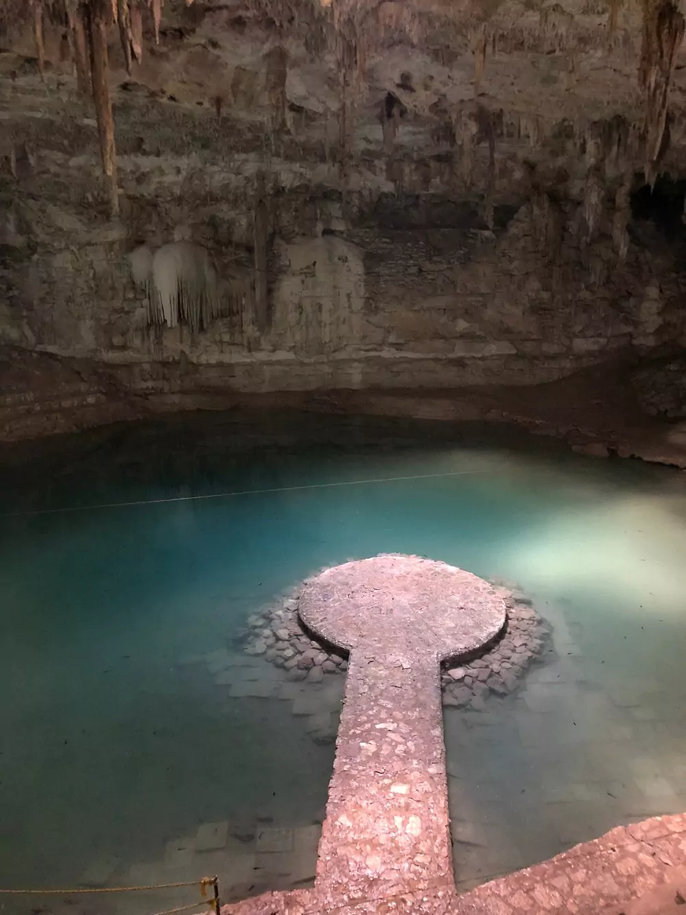 Cenote Suytun in Mexico, featuring a circular stone platform in the center of a shallow, crystal-clear pool, surrounded by high limestone walls with a dramatic beam of light shining through an opening above.