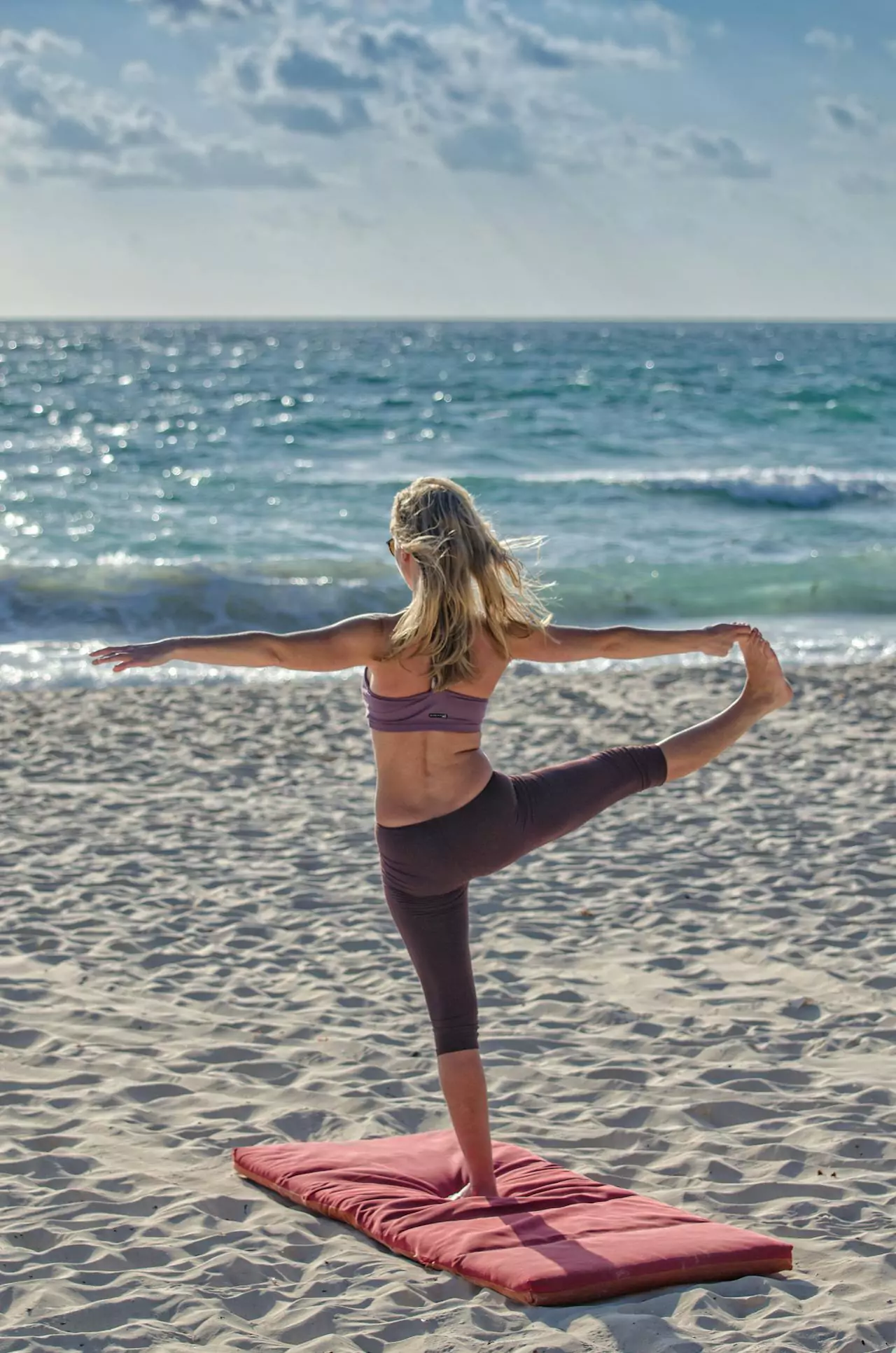 A woman doing yoga on a beach, balancing on one leg with ocean waves in the background.