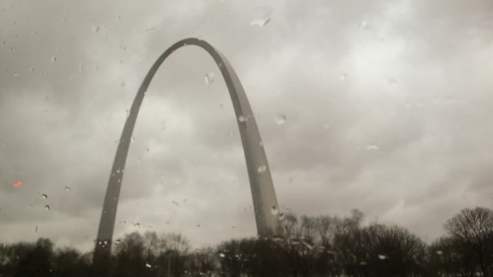 The iconic St. Louis Arch towering into a gray, rainy sky with wet pavement and reflections on the ground. The overcast weather creates a moody atmosphere around the famous landmark.