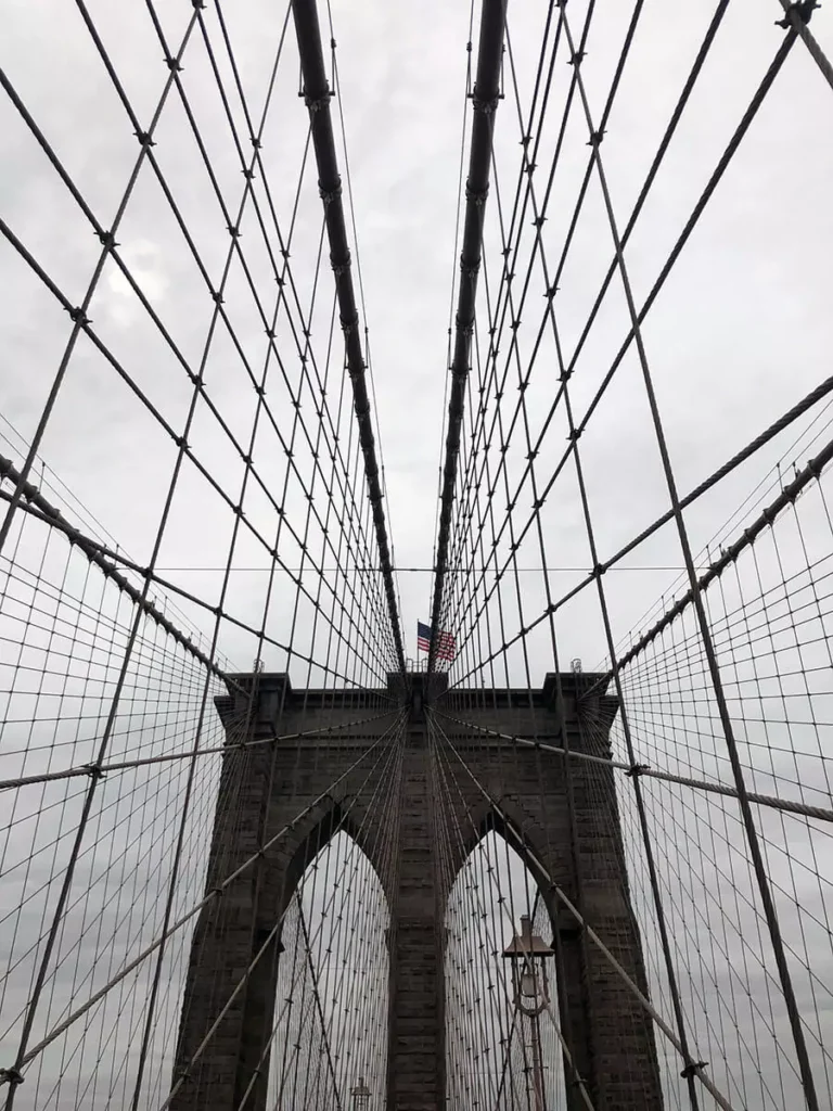 Brooklyn Bridge shrouded in thick fog, with its suspension cables fading into the mist and only the closest stone tower clearly visible. The moody atmosphere softens the usual city skyline, creating a quiet, almost ethereal view.